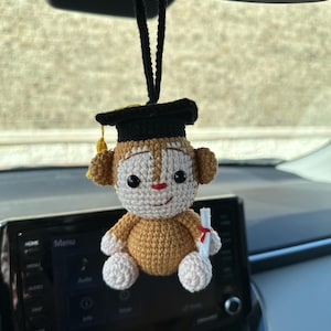 May include: A crocheted monkey wearing a black graduation cap with a yellow tassel, holding a white scroll with a red ribbon. The monkey is hanging from the rearview mirror of a car.