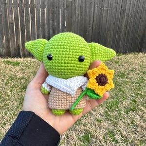May include: A crocheted green figure with large ears, black eyes, and a tan outfit, holding a yellow sunflower with a brown center and green stem. The figure has a white collar and green feet. The background is a wooden fence and grass.
