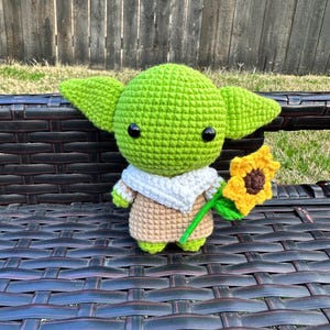 May include: A handmade crochet doll in the likeness of a popular character, featuring large green ears, black eyes, and a tan robe. The doll is holding a yellow sunflower with a green stem. The doll is sitting on a dark brown bench.