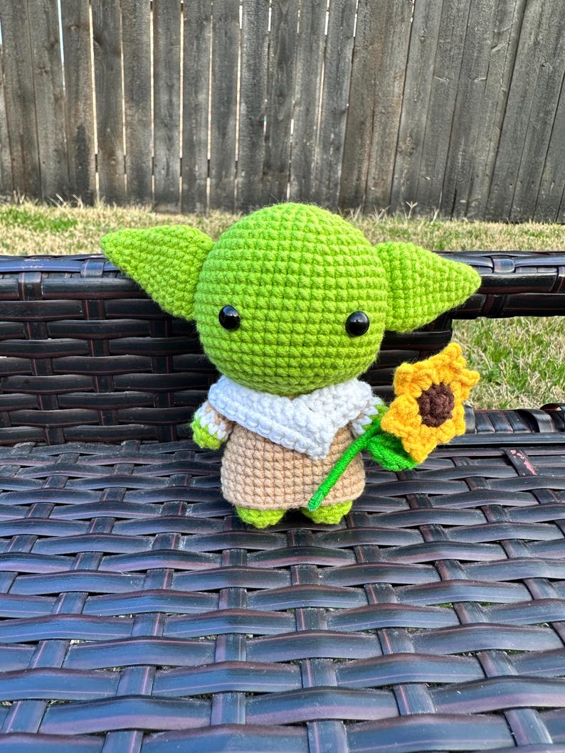 May include: A handmade crochet doll in the likeness of a popular character, featuring large green ears, black eyes, and a tan robe. The doll is holding a yellow sunflower with a green stem. The doll is sitting on a dark brown bench.