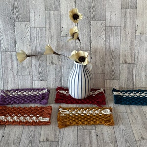May include: Five woven macrame coasters in different colors, including purple, red, orange, and teal, with white accents. The coasters are arranged in a row on a wood floor. A white vase with blue stripes and a dried flower arrangement is in the center of the coasters.