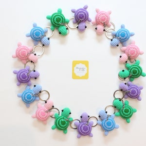 May include: A collection of 14 crocheted turtle keychains in various colors, including green, blue, purple, and pink. The keychains are arranged in a circle on a white background.