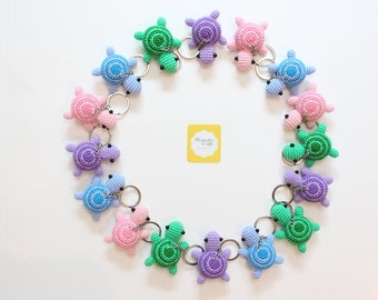 May include: A collection of 14 crocheted turtle keychains in various colors, including green, blue, purple, and pink. The keychains are arranged in a circle on a white background.