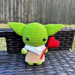 May include: A handmade crochet doll in the likeness of a popular character, featuring large green ears, a tan robe, and a white scarf. The doll is holding a red heart on a stick, perfect for a gift.