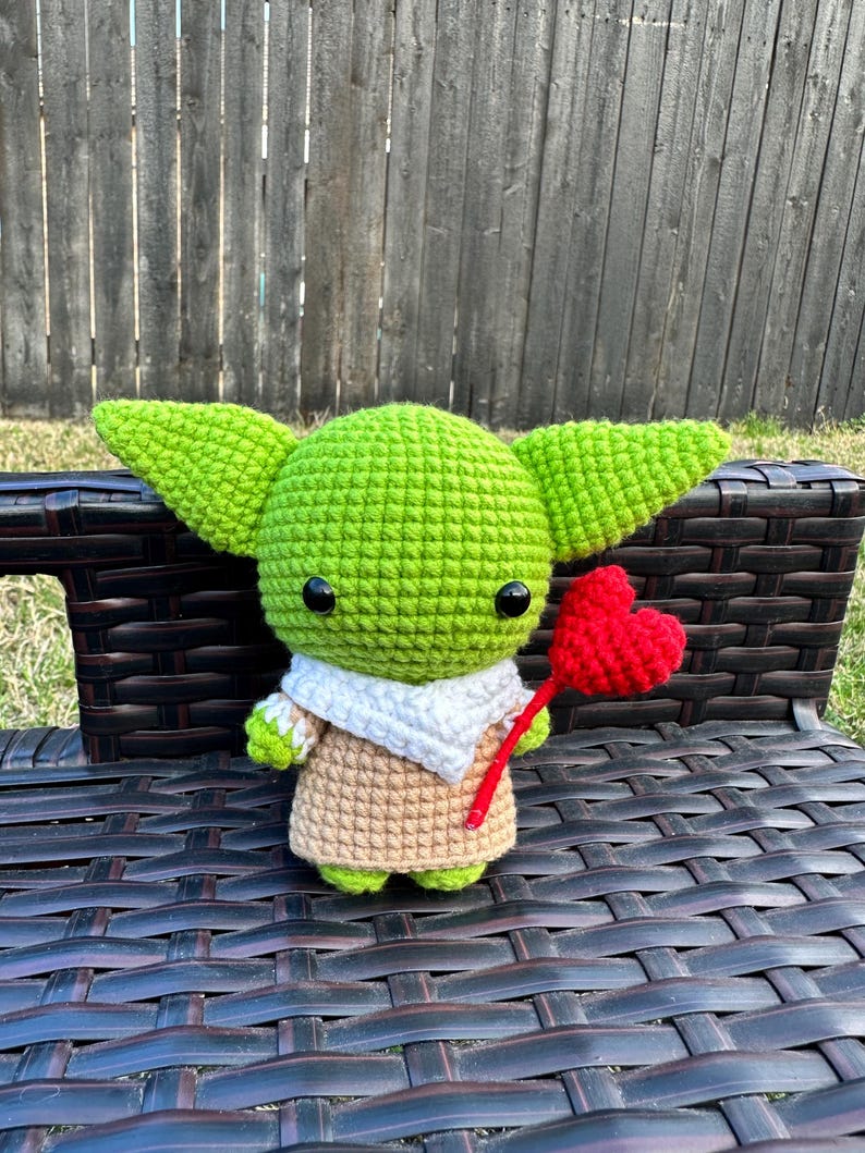 May include: A handmade crochet doll in the likeness of a popular character, featuring large green ears, a tan robe, and a white scarf. The doll is holding a red heart on a stick, perfect for a gift.