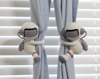 May include: Two white crocheted astronaut-shaped curtain tiebacks with gray visors and red accents holding back blue curtains.