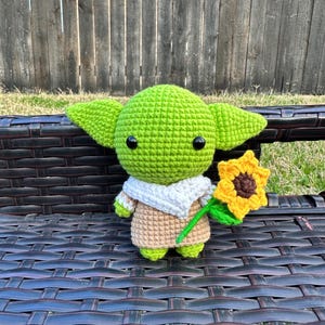 May include: A handmade crochet figure of a green alien-like character with large ears, black eyes, and a tan and white outfit. The figure is holding a yellow sunflower and is sitting on a dark brown bench.