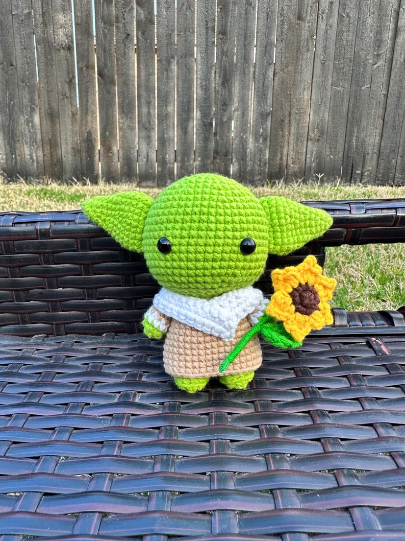 May include: A handmade crochet figure of a green alien-like character with large ears, black eyes, and a tan and white outfit. The figure is holding a yellow sunflower and is sitting on a dark brown bench.
