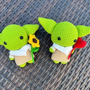May include: Two crocheted green figures with large ears, black eyes, and tan outfits. One holds a yellow sunflower, the other a red heart. Both figures have white collars and are on a brown woven surface.