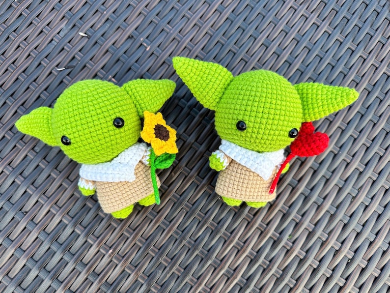 May include: Two crocheted green figures with large ears, black eyes, and tan outfits. One holds a yellow sunflower, the other a red heart. Both figures have white collars and are on a brown woven surface.