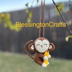 May include: A brown crocheted monkey holding a yellow banana. The monkey has white eyes and a pink nose. The monkey is hanging from a branch.