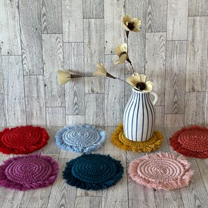 May include: Seven colorful round crocheted coasters with fringe, arranged in a semi-circle on a woodgrain surface. A vase with white and blue stripes holds a bouquet of white flowers.