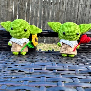 May include: Two crocheted green figures with large ears, black eyes, and tan outfits. One holds a sunflower, the other a red heart. They are sitting on a dark brown woven surface, with a wooden fence in the background.