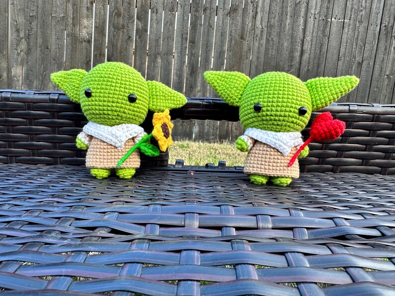 May include: Two crocheted green figures with large ears, black eyes, and tan outfits. One holds a sunflower, the other a red heart. They are sitting on a dark brown woven surface, with a wooden fence in the background.