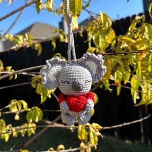 May include: A gray crocheted koala holding a red heart. The koala is hanging from a branch of a tree with yellow leaves.