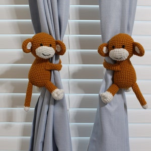 May include: Two brown crocheted monkey curtain tiebacks with white faces and white arms and legs. The monkeys are holding onto light blue curtains.