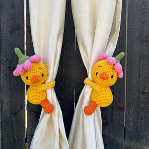 May include: Two yellow crochet duck curtain tiebacks, each featuring a small orange beak, black eyes, and a pink flower-shaped hat with a green stem. The duck figures are designed to wrap around and hold back light-colored fabric curtains.