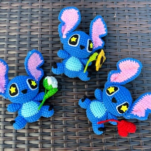 May include: Three handmade blue crochet figures with large pink-lined ears, yellow star-shaped eyes, and light blue bellies. One figure holds a green and white item, another holds a yellow item, and the third holds a red item. The figures are arranged on a brown woven surface.