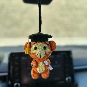 May include: A crocheted orange and yellow Tigger wearing a black graduation cap and holding a diploma. The Tigger is hanging from a rearview mirror in a car.