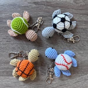 May include: Four crocheted turtle keychains, each designed to resemble a different sports ball: tennis, soccer, basketball, and baseball. The turtles are in green, black and white, orange, and blue, with silver-colored key rings.