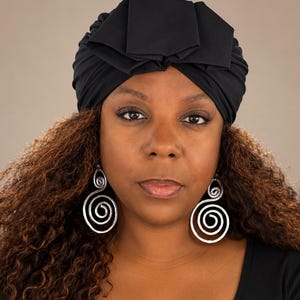 May include: A solid black fabric turban featuring a structured, folded knot detail at the centre front. The headwear is paired with large, silver-toned metal earrings that showcase a prominent, hammered spiral design hanging from the earlobes.