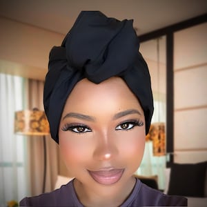 May include: A black head wrap tied in a knot on top of the head. The person in the image has brown eyes and is wearing makeup. The background is blurred, showing a room with a bed and a lamp.