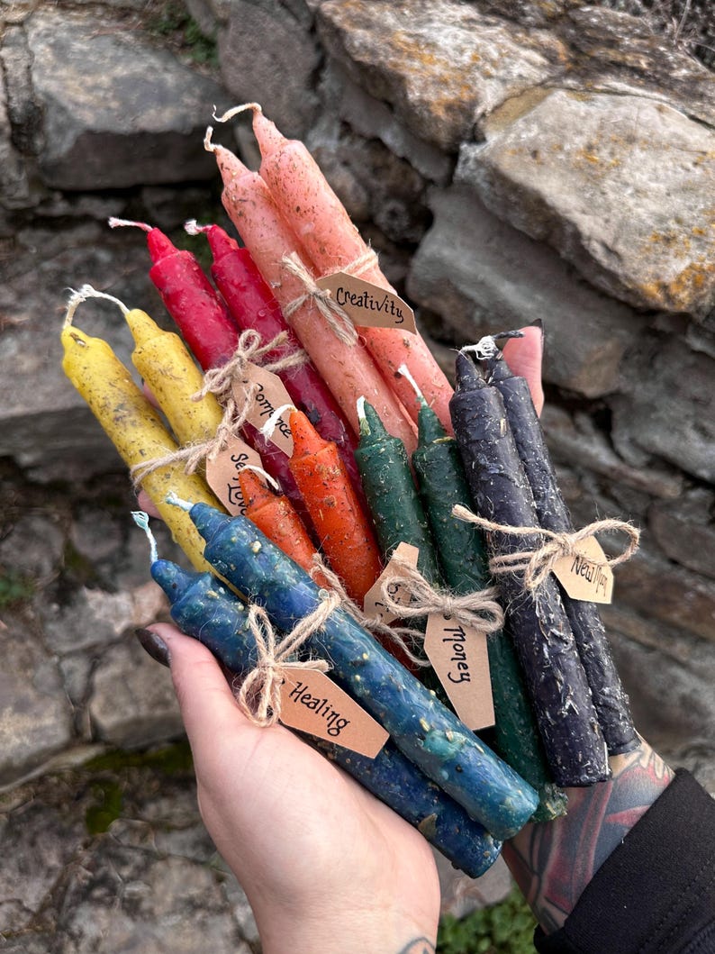 May include: A collection of colorful, handmade spell candles in various shades, including pink, red, yellow, orange, green, blue, and purple. Each candle is adorned with a small tag tied with twine, labeled with words like "Creativity" and "Healing".