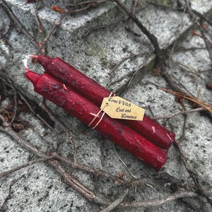 May include: Two deep red, hand-poured spell candles tied together with twine. A small tag reads "Coma Vvitch Lust and Romance". The candles rest on a textured stone surface with vines.