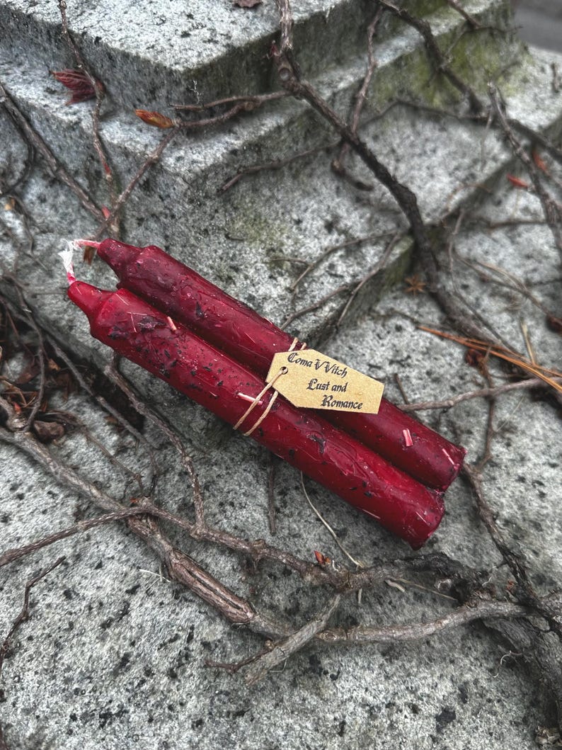 May include: Two deep red, hand-poured spell candles tied together with twine. A small tag reads "Coma Vvitch Lust and Romance". The candles rest on a textured stone surface with vines.