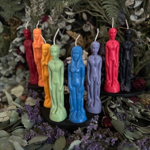 May include: A set of nine human-shaped candles in various colors, including red, orange, yellow, green, blue, purple, and black. The candles are arranged in a semi-circle on a black surface.