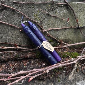 May include: Two dark purple candles with a textured surface, resting on a weathered stone surface with vines. A small tag tied to one candle reads "Emma V Vitch Manifest". The candles are a deep shade of purple.