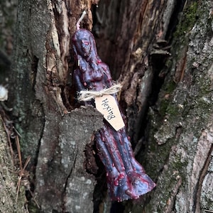 Könnte beinhalten: Eine dunkelviolette und rote Kerze in Form einer menschlichen Figur mit einem Etikett, auf dem "Hexing" steht.