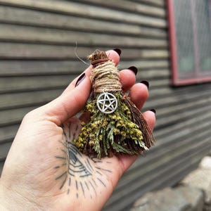 Op de afbeelding: Een kleine, handgemaakte bezem met gedroogde kruiden en een zilveren pentagrambedel. De bezem is omwikkeld met touw en heeft een bosje gedroogde gele en groene bloemen. De achtergrond is een verweerde houten muur.