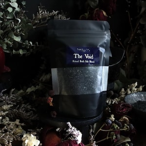 May include: A black pouch labeled "The Void" containing a ritual bath salt blend. The pouch is partially transparent, revealing the salt. The image is set against a dark background with dried flowers and a pomegranate.