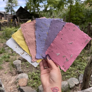 May include: A hand holds a fan of handmade paper sheets in various colors: white, yellow, pink, purple, and rose. The paper has a textured surface with visible inclusions. The background features a wooden fence and greenery.
