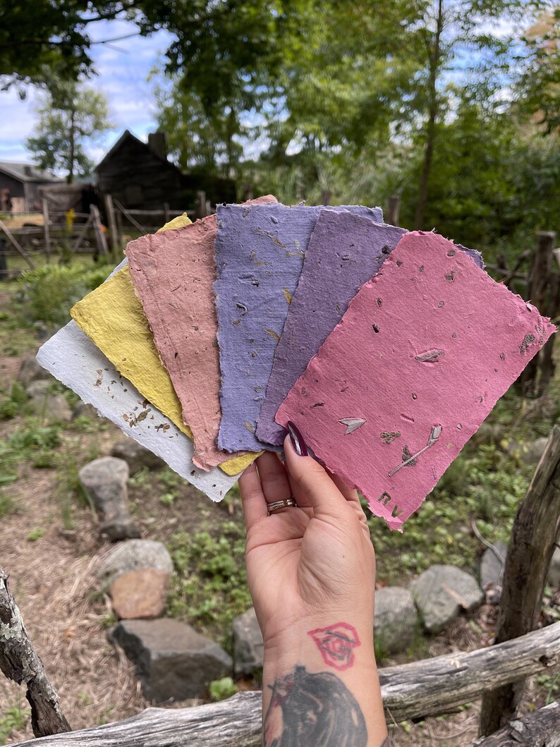 May include: A hand holds a fan of handmade paper sheets in various colors: white, yellow, pink, purple, and rose. The paper has a textured surface with visible inclusions. The background features a wooden fence and greenery.