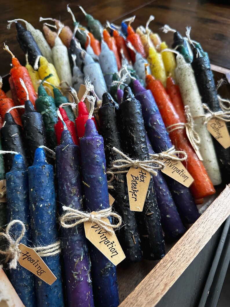 K&ouml;nnte beinhalten: Eine Sammlung bunter, handgerollter und beschrifteter Kerzen in einer Holzkiste. Die Etiketten beinhalten "Hex Breaker", "Divination", "Healing" und "Cord Cutting".