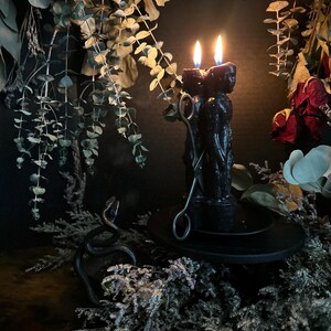 May include: A black candle with two lit flames, featuring a sculpted design, sits on a black plate. A metal snake sculpture and candle snuffer are nearby. The scene is decorated with dried foliage and flowers, creating a gothic or mystical aesthetic.