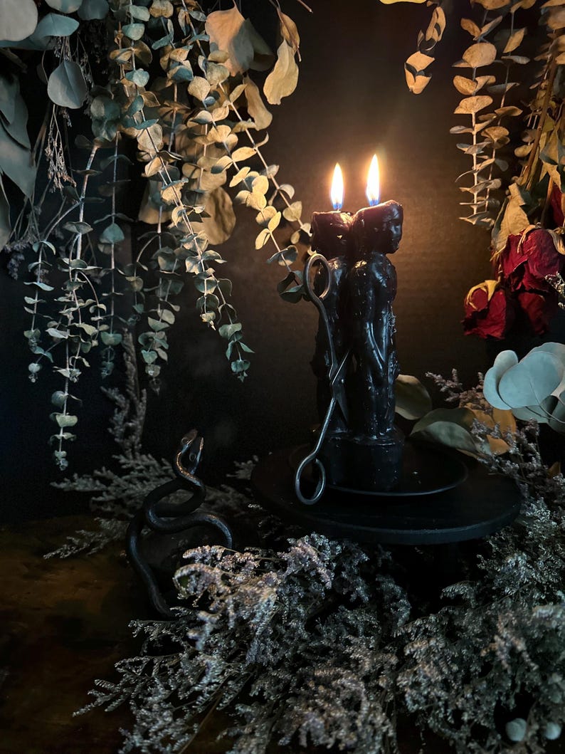 May include: A black candle with two lit flames, featuring a sculpted design, sits on a black plate. A metal snake sculpture and candle snuffer are nearby. The scene is decorated with dried foliage and flowers, creating a gothic or mystical aesthetic.