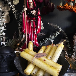 Może przedstawiać: Zestaw żółtych świec w ciemnej misce, z naklejką "Come Witch Santa Muerte". Czerwona figura szkieletu z kosą stoi za świecami. Tło jest ciemne.