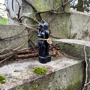 May include: A black candle in the shape of two figures, tied with twine and a small tag. The candle is set against a weathered stone surface with moss and vines. The tag reads "Love Hate Separation".