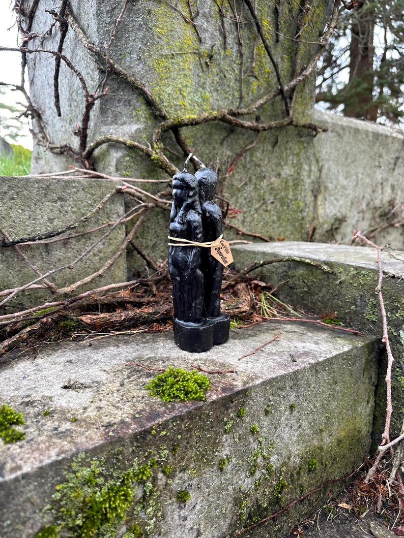 May include: A black candle in the shape of two figures, tied with twine and a small tag. The candle is set against a weathered stone surface with moss and vines. The tag reads "Love Hate Separation".