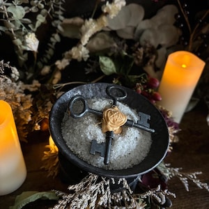 May include: A still-life arrangement featuring three antique keys arranged in a cross formation, centered on a bed of white crystals within a dark, round bowl. A gold-colored pendant with a star design adorns the keys. Lit candles and dried flowers create a mystical ambiance.