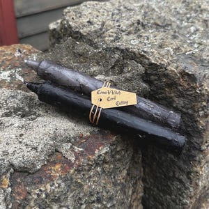 Può includere: Due candele rituali, una nera e una grigia, poggiano su una superficie rocciosa strutturata. Un piccolo cartellino legato con spago recita "Coma Witch Cord Cutting". Le candele sono cilindriche e sembrano fatte a mano.