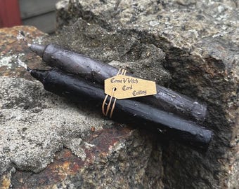 May include: Two ritual candles, one black and one gray, rest on a textured rock surface. A small tag tied with twine reads "Coma Witch Cord Cutting". The candles are cylindrical and appear handmade, suggesting a focus on spiritual practices.