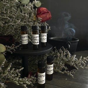 Op de afbeelding: Vijf kleine glazen flesjes met zwarte dopjes en witte etiketten. De etiketten hebben handgeschreven tekst die "ComaVV", "Honey Magic", "Anti Festive", "The Lovers" en "Protection" luidt. De flesjes zijn gerangschikt op een zwarte standaard met een zwarte achtergrond.