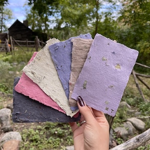 May include: A hand holds a fan of handmade paper sheets in various colors: dark gray, pink, beige, blue, and lavender. The paper has a rough texture and visible plant fibers. The lavender sheet has the initials "M T" printed on it.