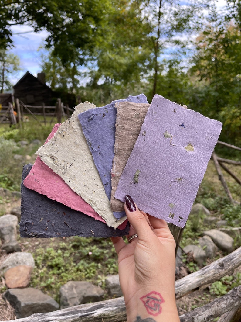 May include: A hand holds a fan of handmade paper sheets in various colors: dark gray, pink, beige, blue, and lavender. The paper has a rough texture and visible plant fibers. The lavender sheet has the initials "M T" printed on it.