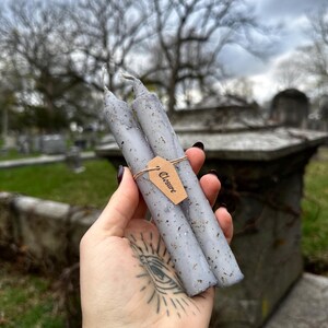 May include: Two gray spell candles with flecks of herbs are held together with a brown tag that reads "Closure". The candles are cylindrical and have a rustic, handmade appearance. The background is a blurred outdoor scene with trees and a cloudy sky.