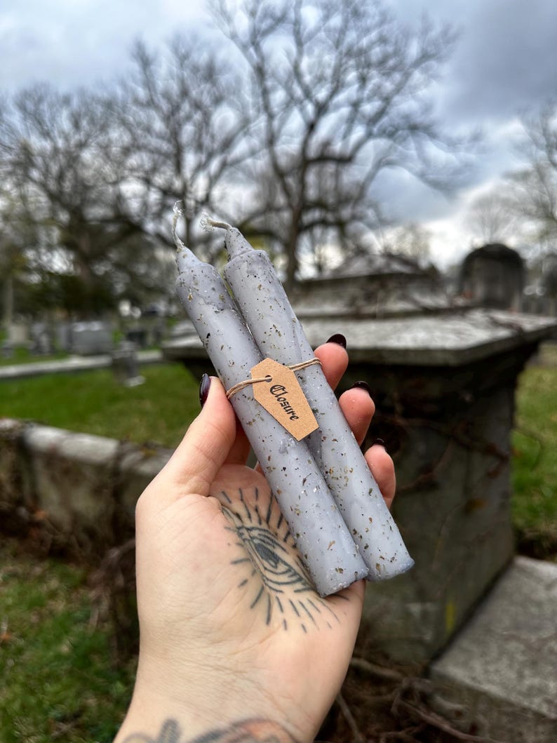 May include: Two gray spell candles with flecks of herbs are held together with a brown tag that reads "Closure". The candles are cylindrical and have a rustic, handmade appearance. The background is a blurred outdoor scene with trees and a cloudy sky.